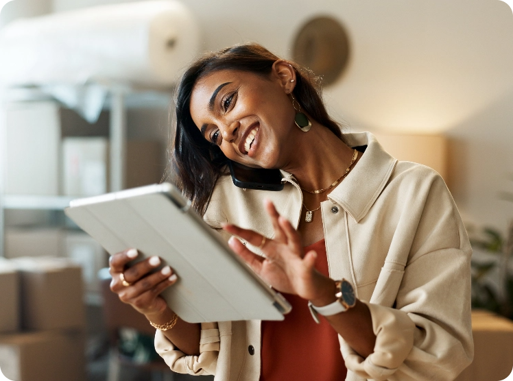 Smiling woman swipes on a tablet screen while holding a phone between her ear and shoulder